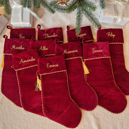 Eksklusiv rød julestrømpe i fløyel med personlig brodert navn “Thomas”. Dekorert med elegant gullfrynse og hengende på juletre med julelys og pynt. Eventyrprint.no – perfekt personlig julestrømpe i fløyel til jul. Mange julestrømper