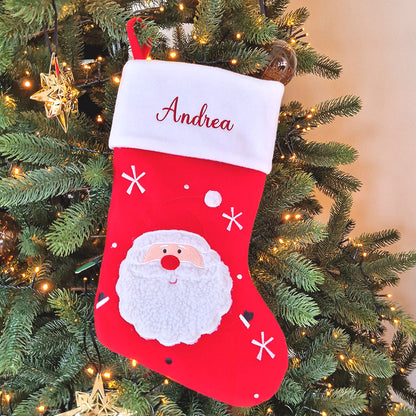 Rød julestrømpe med hvit mansjett og brodert navn, dekorert med stor julenisse og snøfnuggmotiv. Andrea på tre