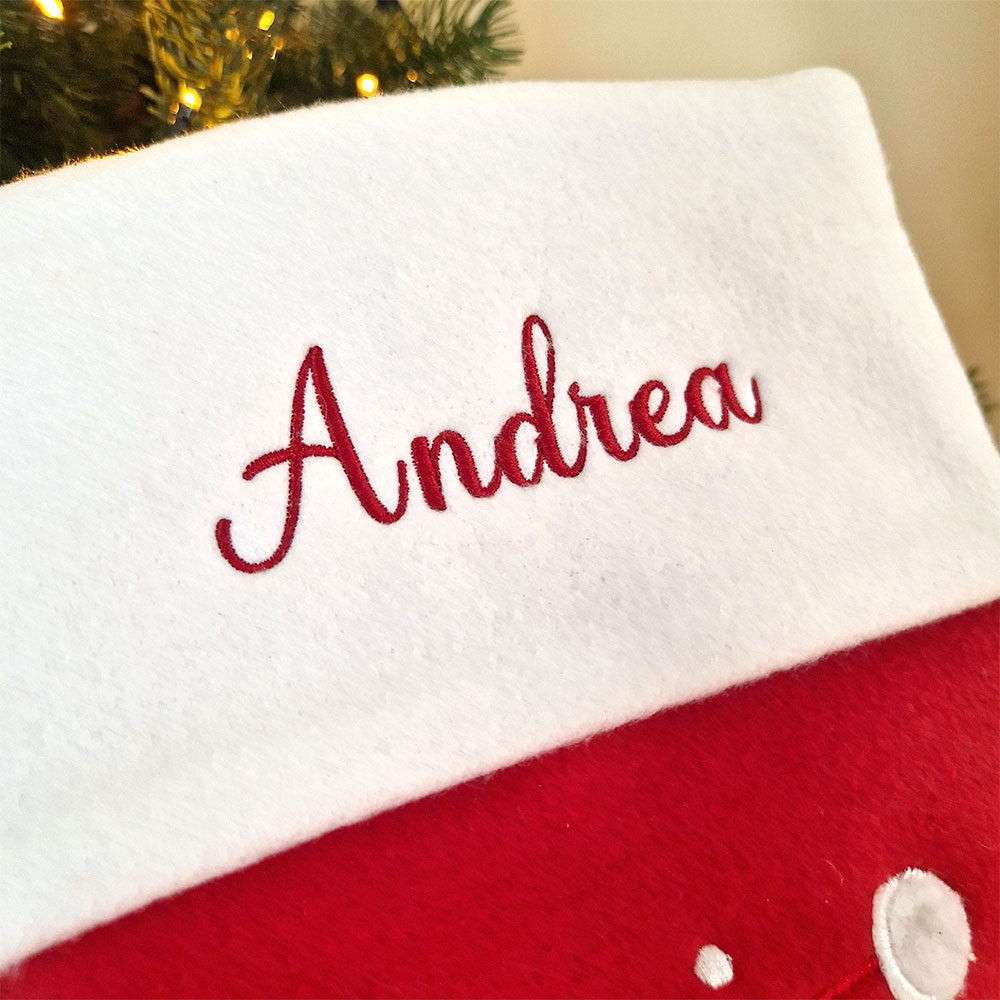 Rød julestrømpe med hvit mansjett og brodert navn, dekorert med stor julenisse og snøfnuggmotiv. Nærbilde av navn