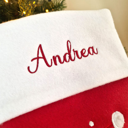 Rød julestrømpe med hvit mansjett og brodert navn, dekorert med stor julenisse og snøfnuggmotiv. Nærbilde av navn
