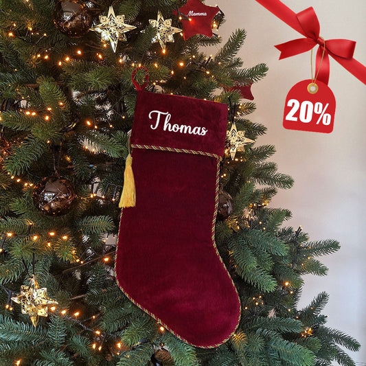 Eksklusiv rød julestrømpe i fløyel med personlig brodert navn “Thomas”. Dekorert med elegant gullfrynse og hengende på juletre med julelys og pynt. Nå med 20 % rabatt hos Eventyrprint.no – perfekt personlig julestrømpe i fløyel til jul.