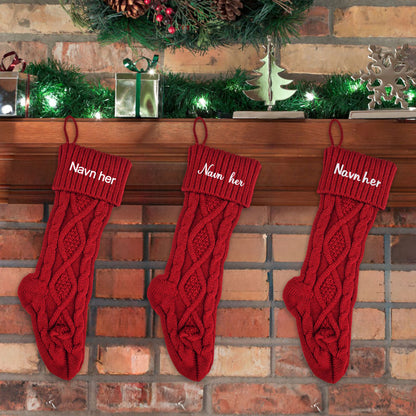 Tre røde strikkede julestrømper med flettemønster henger på peisen, hver med brodert personlig navn.